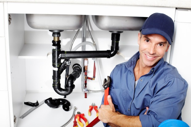A plumber wearing a blue cap and shirt sits in front of an open cabinet under a kitchen sink, holding a wrench with plumbing tools and pipes visible.
